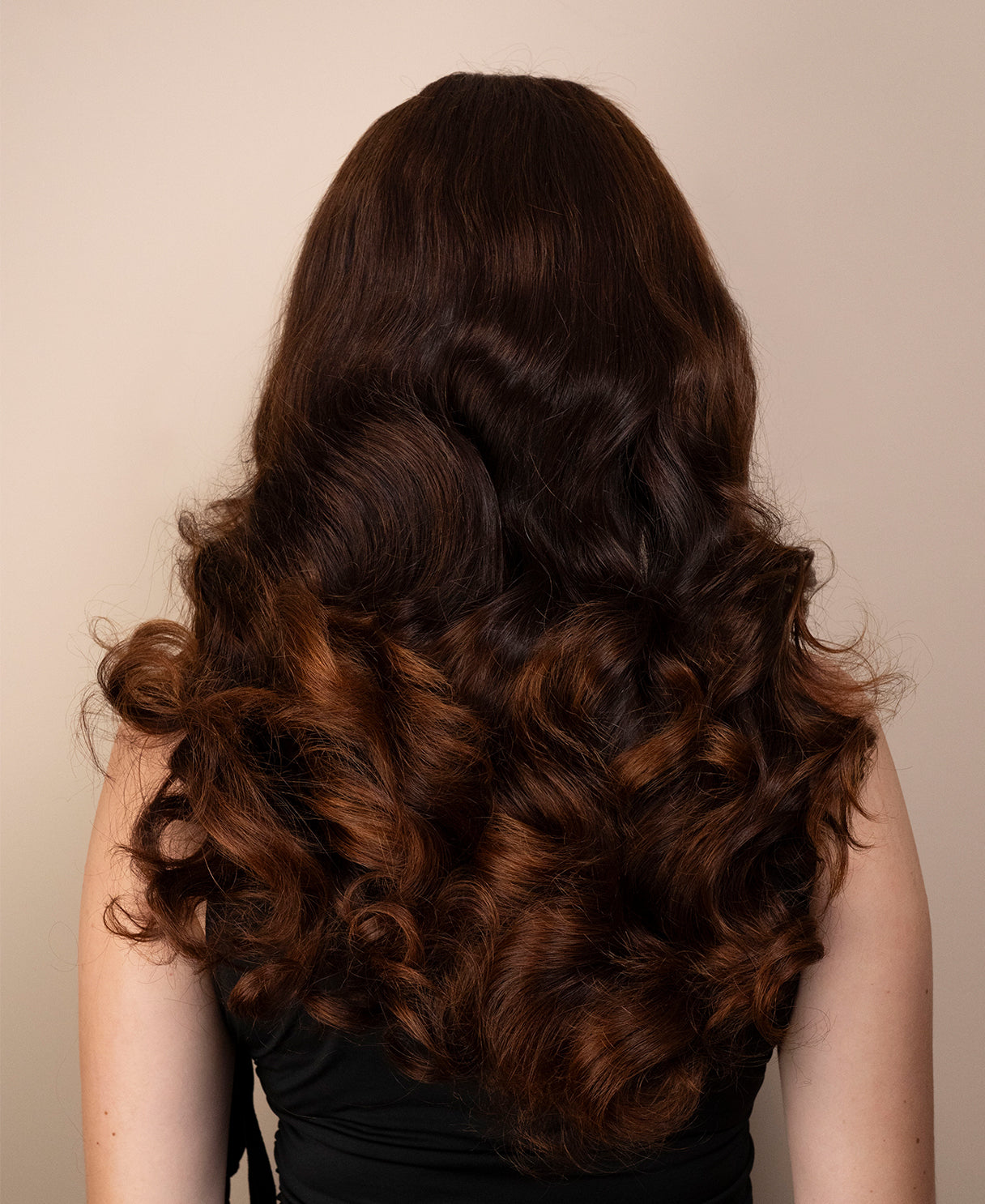 Woman with long, wavy brown hair wig wearing a black top against a beige background