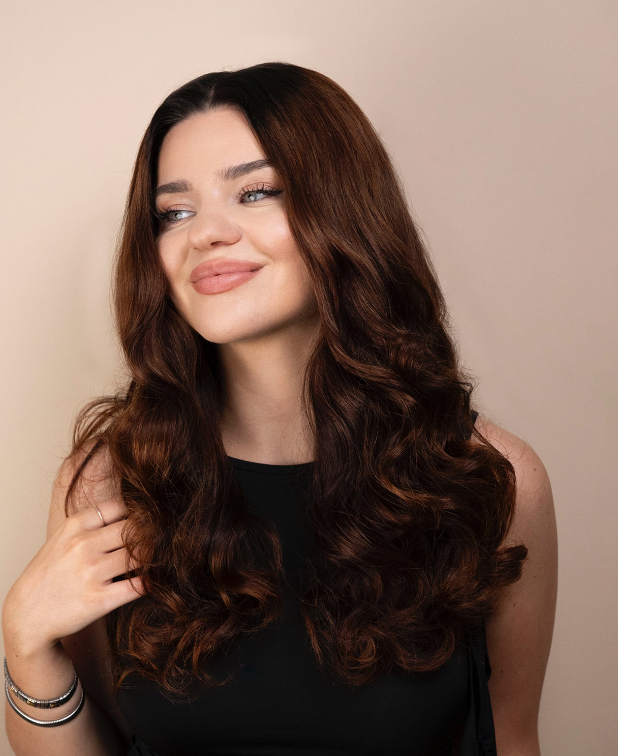 Woman with long, wavy brown hair wig wearing a black top against a beige background
