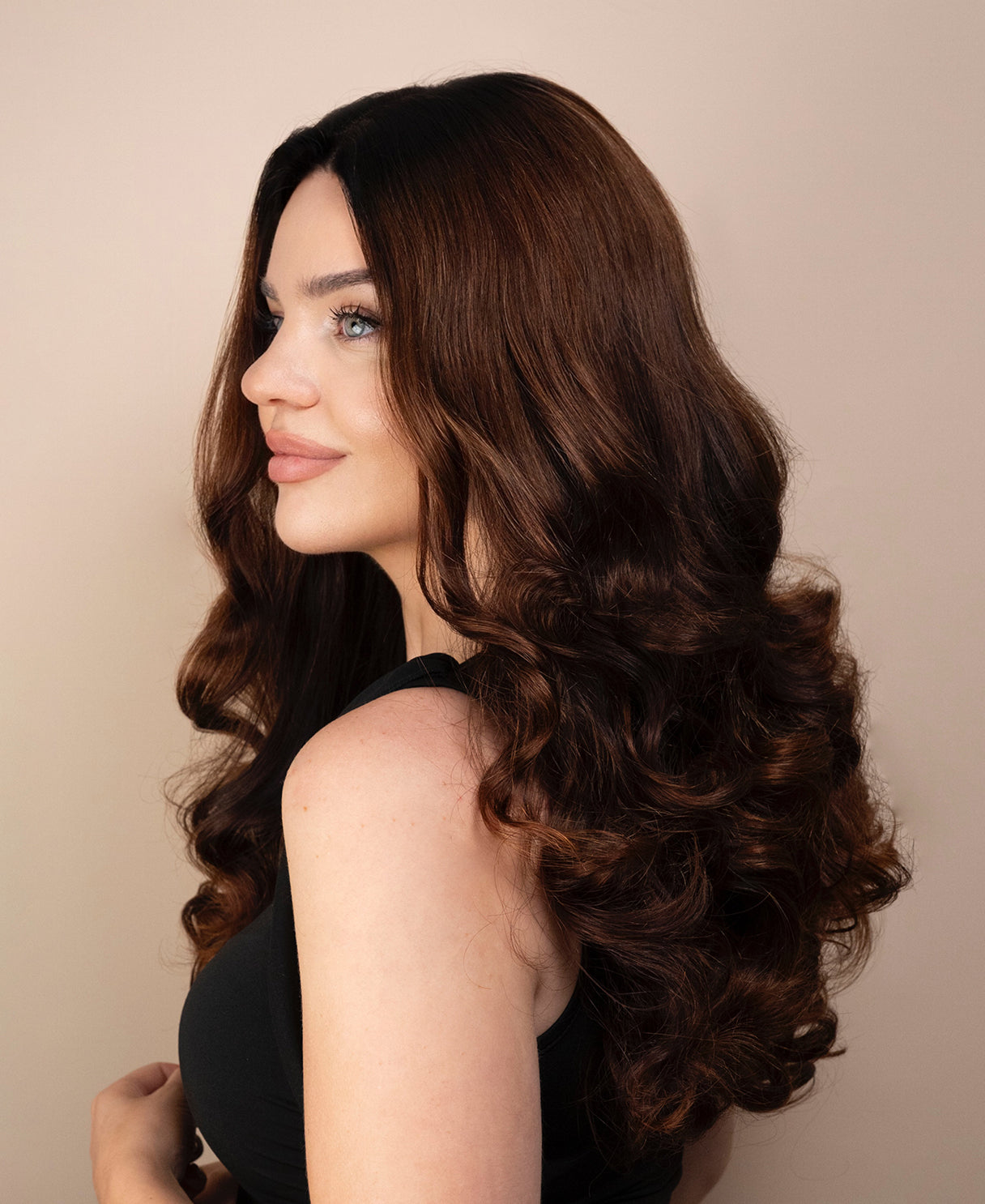 Woman with long, wavy brown hair wig wearing a black top against a beige background