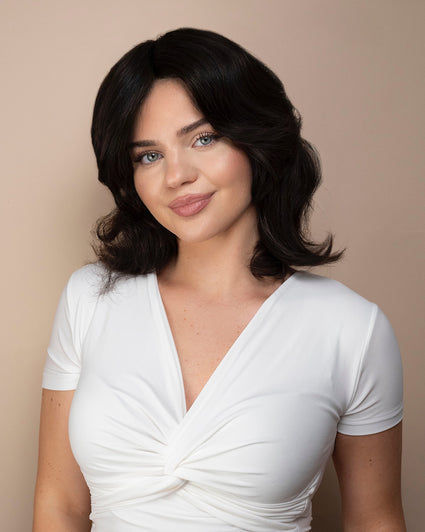 Woman with shoulder-length dark wig hair wearing a white top against a beige background