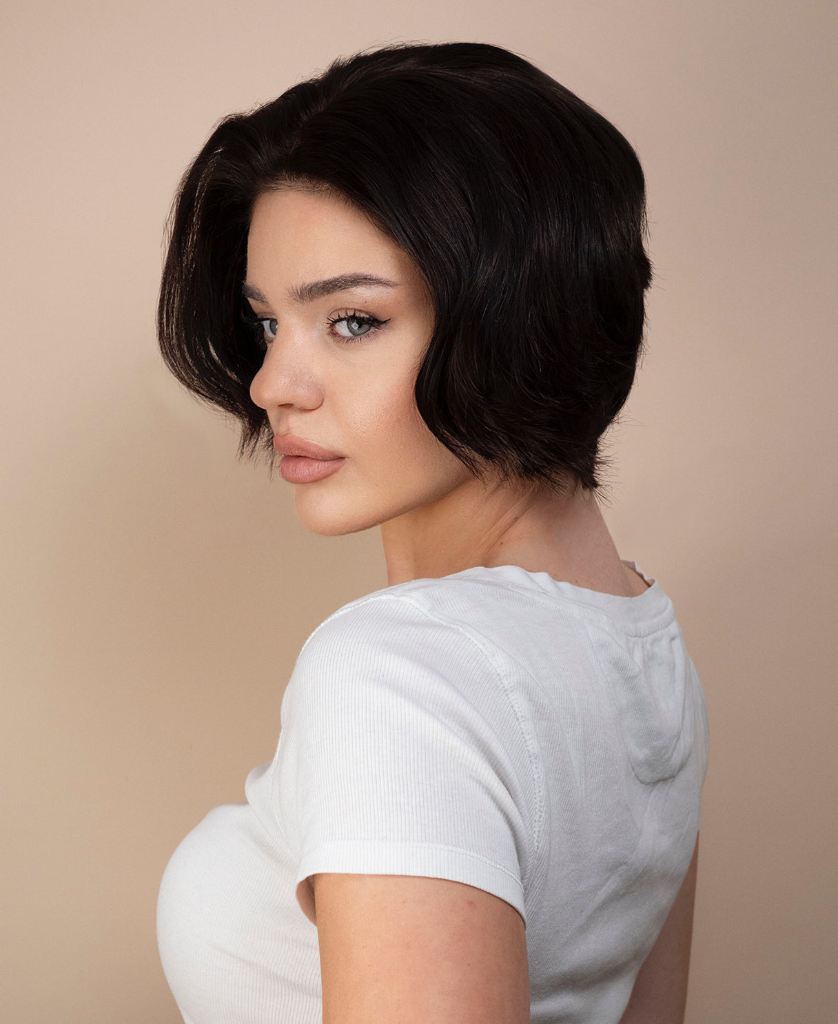 Woman with a short black bob haircut wearing a white t-shirt against a beige background