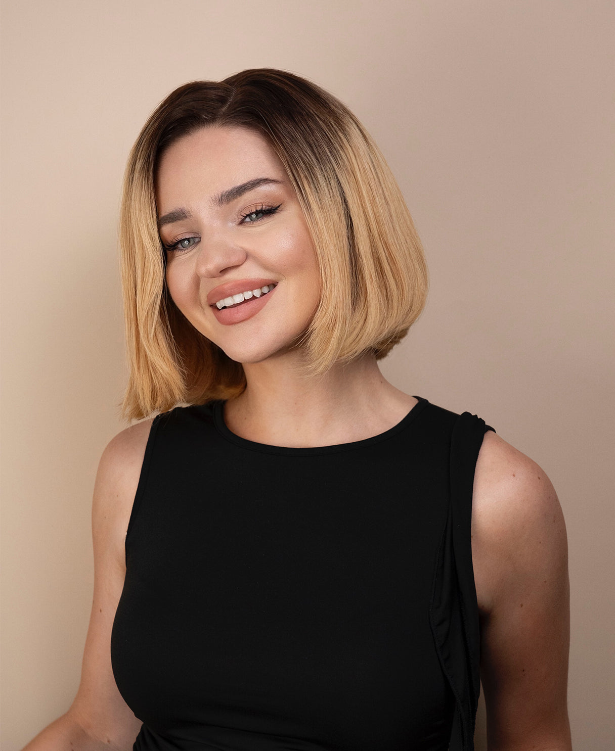 Woman with a stylish blonde haircut wig wearing a black sleeveless top against a beige background