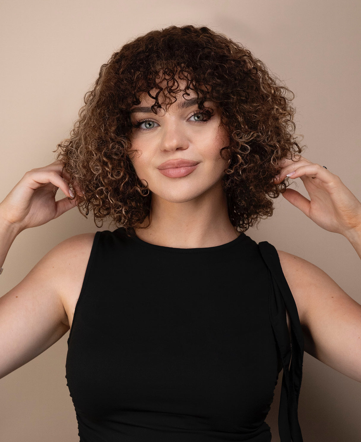 Woman with curly wig wearing a black top against a beige background