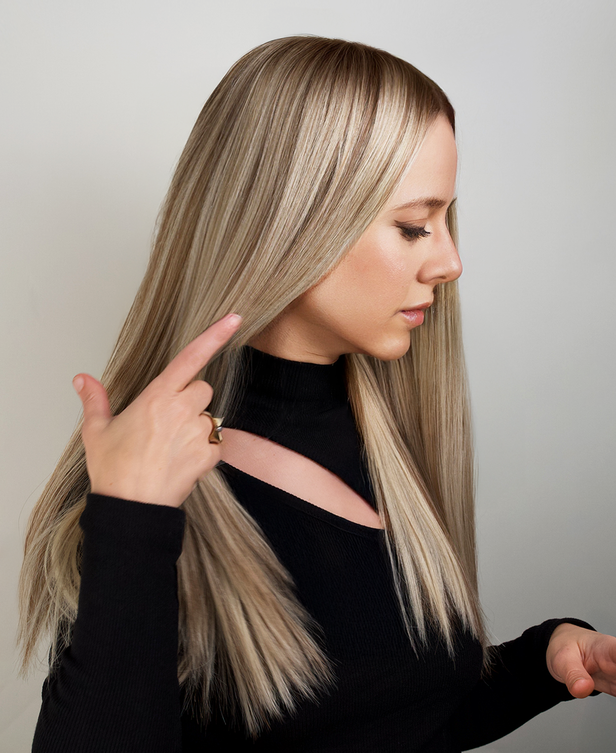 Woman with long blonde hair wig pointing to her hair, wearing a black top.